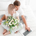 Paar sitzt im Bett, sie hält einen Strauß weißer Rosen in den Händen, er tippt etwas in einen Laptop.