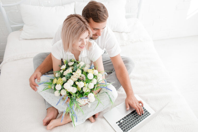 Paar sitzt im Bett, sie hält einen Strauß weißer Rosen in den Händen, er tippt etwas in einen Laptop.