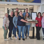 Mehrere Personen stehen in einem Raum zum Gruppenfoto versammelt, im Hintergrund ist ein Anmeldeterminal zu sehen.