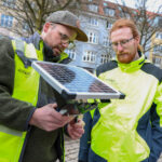 Zwei Männer in neongelber Warnkleidung auf einem städtischen Platz, sie betrachten ein kleines Solarmodul