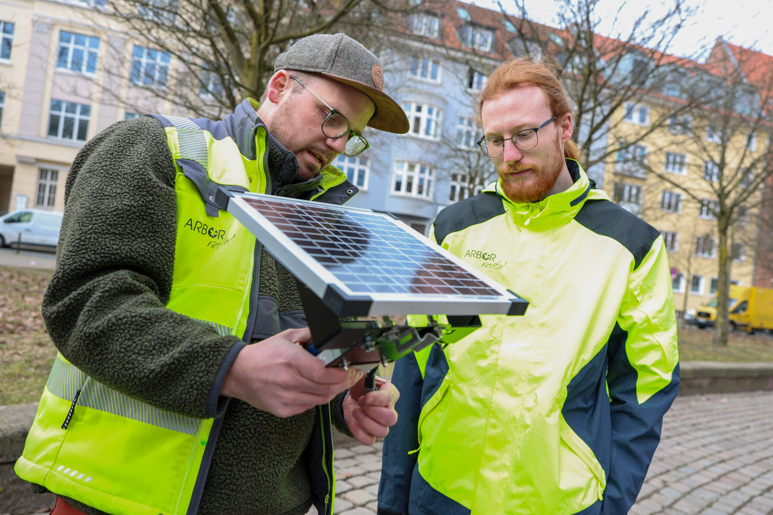 Zwei Männer in neongelber Warnkleidung auf einem städtischen Platz, sie betrachten ein kleines Solarmodul