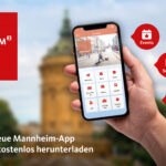 "Ganz Mannheim in deiner Hand" - Logo der Mannheimer City App, hochgehaltenes Handy mit der App und im Hintergrund ein städtischer Platz mit historischem Turm.
