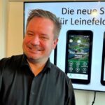 Bürgermeister Christian Zwingmann hält ein Smartphone in der Hand, auf dem die Leinefelde-Worbis-App zu sehen ist, im Hintergrund sind auf einem Display weitere App-Ansichten zu sehen.