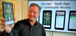 Bürgermeister Christian Zwingmann hält ein Smartphone in der Hand, auf dem die Leinefelde-Worbis-App zu sehen ist, im Hintergrund sind auf einem Display weitere App-Ansichten zu sehen.