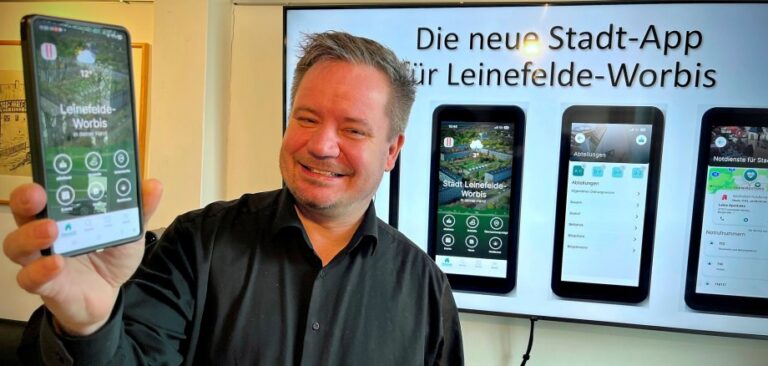 Bürgermeister Christian Zwingmann hält ein Smartphone in der Hand, auf dem die Leinefelde-Worbis-App zu sehen ist, im Hintergrund sind auf einem Display weitere App-Ansichten zu sehen.