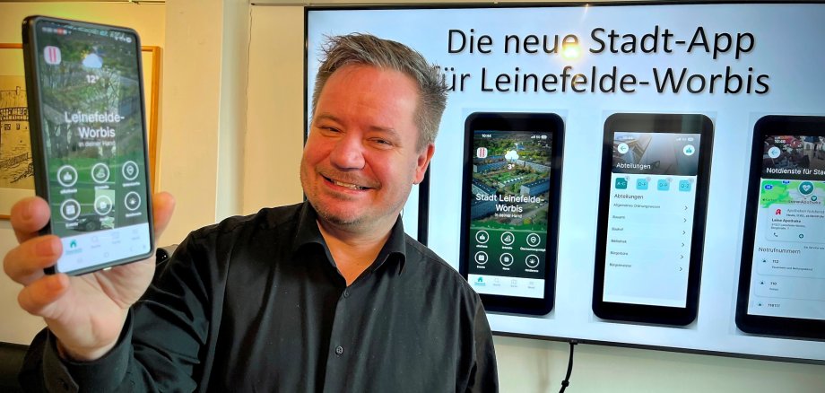 Bürgermeister Christian Zwingmann hält ein Smartphone in der Hand, auf dem die Leinefelde-Worbis-App zu sehen ist, im Hintergrund sind auf einem Display weitere App-Ansichten zu sehen.