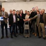 Mehrere Personen stehen in einem Raum zum Gruppenfoto versammelt. Zwei von ihnen betätigen einen symbolischen Startknopf, eine dritte hält einen aufgeklappten Laptop in den Händen auf dem die Startseite der neuen Website zu sehen ist.