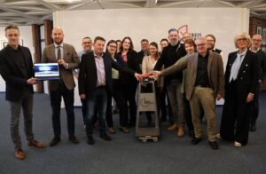 Mehrere Personen stehen in einem Raum zum Gruppenfoto versammelt. Zwei von ihnen betätigen einen symbolischen Startknopf, eine dritte hält einen aufgeklappten Laptop in den Händen auf dem die Startseite der neuen Website zu sehen ist.