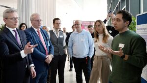 Männer in Anzügen (darunter Bundesminister Wildberger) und junge Menschen in Smart Casual-Kleidung stehen in einem Konferenzraum herum und reden miteinander.
