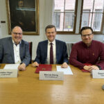 Drei Männer sitzen nebeneinander an einem Tisch. Vor ihnen liegen Vertragsunterlagen.