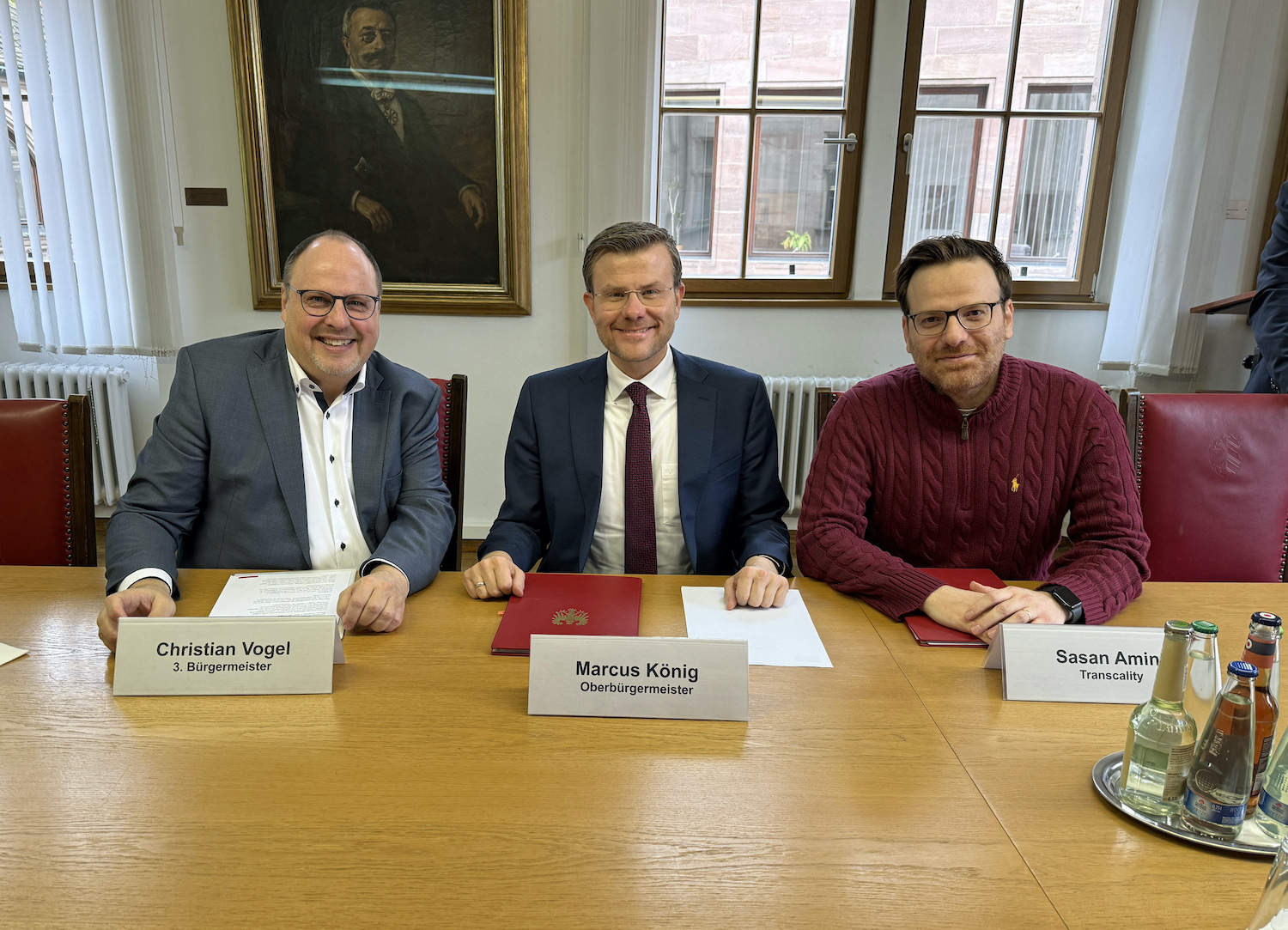 Drei Männer sitzen nebeneinander an einem Tisch. Vor ihnen liegen Vertragsunterlagen.