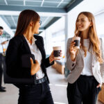 zwei weibliche Mitarbeitende mit Kaffeebechern in der Hand unterhalten sich während sie den Büroflur entlanglaufen