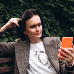 Ein Mädchen in einer braunen Jacke sitzt auf einer Bank und schaut oder liest etwas auf einem Smartphone.