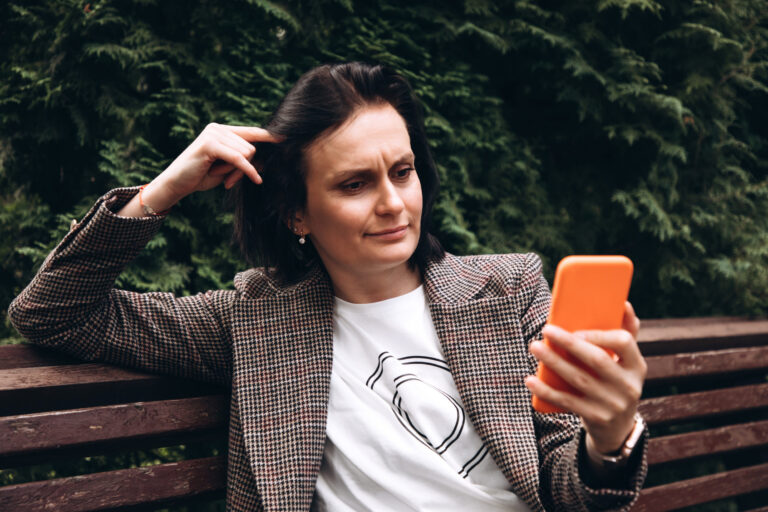 Ein Mädchen in einer braunen Jacke sitzt auf einer Bank und schaut oder liest etwas auf einem Smartphone.