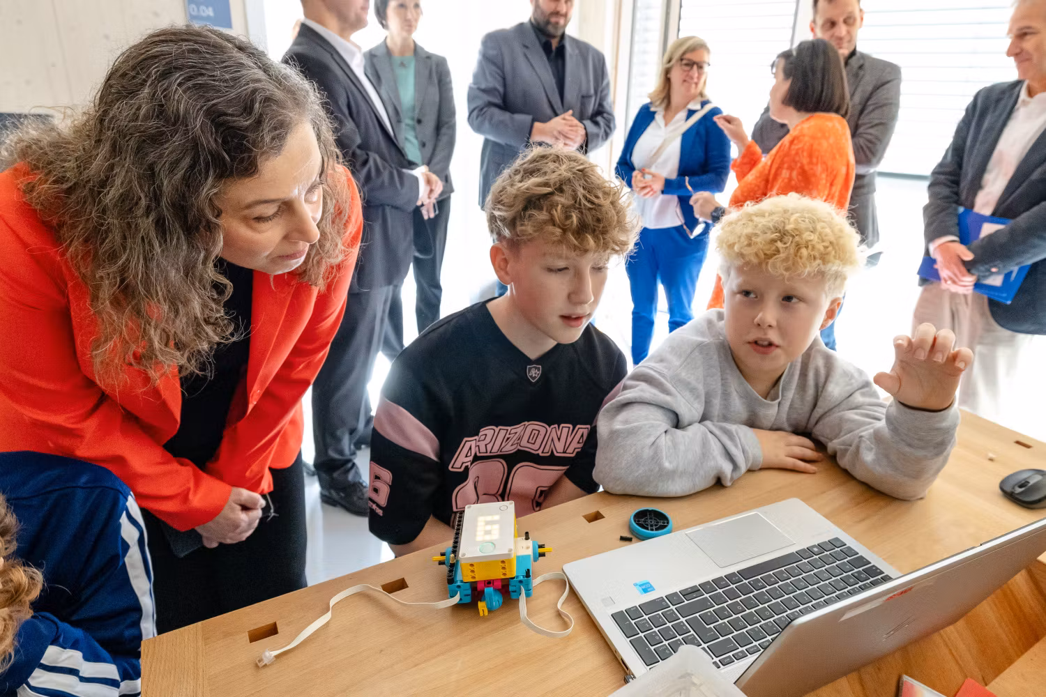 Klassenzimmer-Kulisse: Eine Frau mit langen Locken (Maria Borelli) beugt sich über den Schultisch von zwei Grundschülern, die vor einem Laptop sitzen.