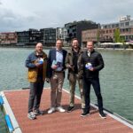Vier Männer auf einem Steg, im Hintergrund Wasserfläche und frisch renovierte bzw. neu gebaute mehrgeschossige Häuser.