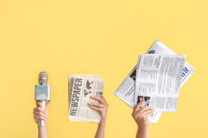 Drei weibliche Hände recken ein Mikrofon und verschiedene Print-Zeitungen hoch; gelber Hintergrund