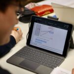 Blick über die Schulter eines jungen, konzentriert erscheinenden Schülers, der auf seinem Tisch ein Tablet und einige Schulutensilien hat.