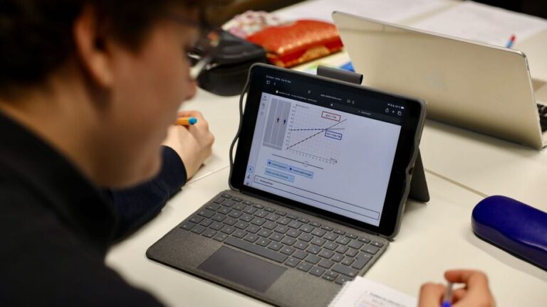 Blick über die Schulter eines jungen, konzentriert erscheinenden Schülers, der auf seinem Tisch ein Tablet und einige Schulutensilien hat.