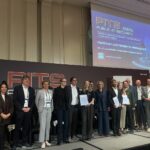 Etwas düster wirkende Bühne mit der obligaten Großprojektion, davor Personen in förmlicher Business-Kleidung in Gruppenfopto-Aufstellung