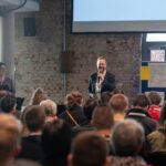 Blick über sitzendes Publikum auf ein Podium mit animiert talkendem Moderator