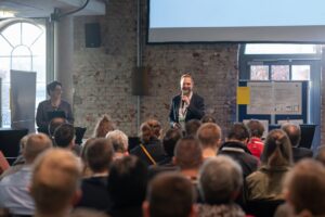 Blick über sitzendes Publikum auf ein Podium mit animiert talkendem Moderator