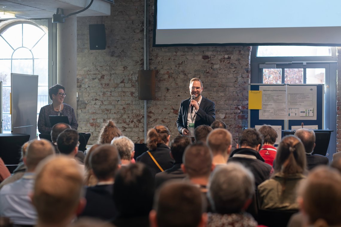 Blick über sitzendes Publikum auf ein Podium mit animiert talkendem Moderator