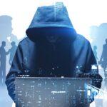 Illustration zum Thema Cybersicherheit: Im Vordergrund sitzt eine dunkel gekleidete Person mit tief ins Gesicht gezogener Kapuze vor einem Laptop. Im Hintergrund sind schemenhafte Silhouetten von Menschen zu sehen, die miteinander sprechen oder arbeiten. Überlagerte digitale Elemente und eine angedeutete Stadtlandschaft in Blau- und Grautönen vermitteln den Eindruck einer vernetzten digitalen Welt und symbolisieren die Bedrohung durch Cyberangriffe auf Organisationen und Verwaltungen.