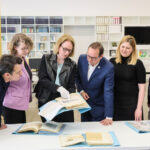 Personengruppe steht um einen Tisch herum und betrachtet Papiere und Kataloge, im Hintergrund Fotos an Stellwänden.