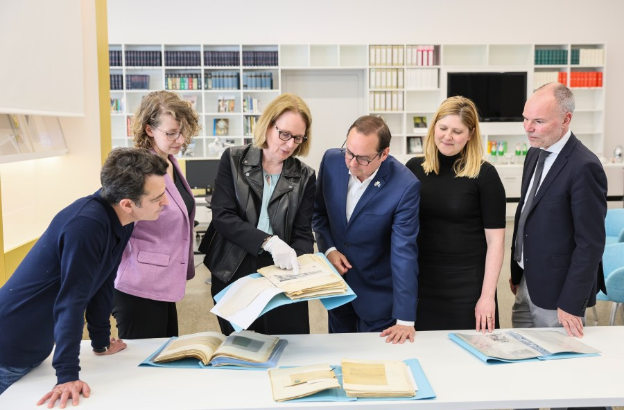 Personengruppe steht um einen Tisch herum und betrachtet Papiere und Kataloge, im Hintergrund Fotos an Stellwänden.