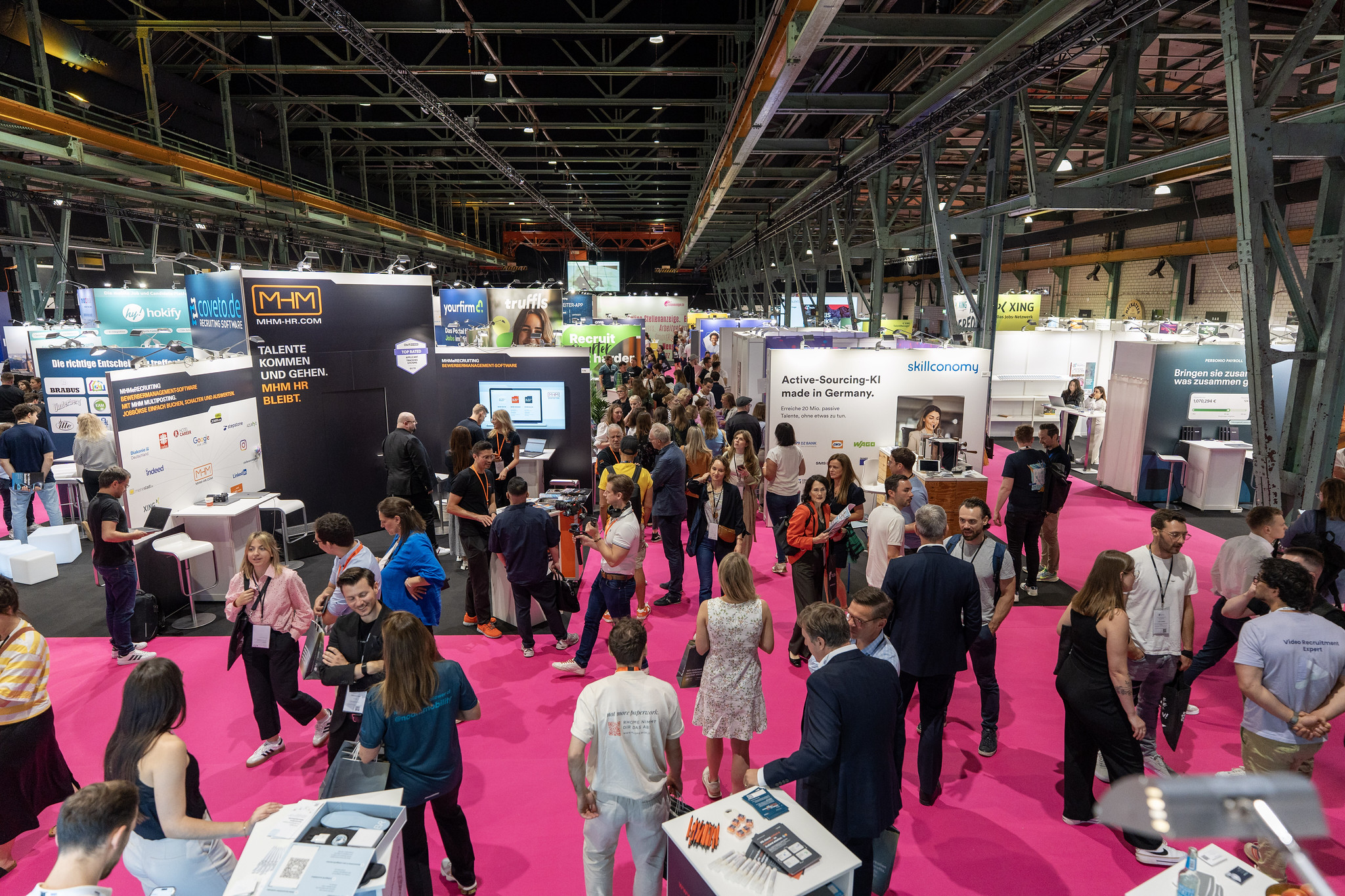 Blick in eine belebte Messehalle mit pinkfarbenem Bodenbelag