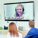 Zwie Personen in einem Meetingraum, auf einem Monitor sind weitere Teilnehmer per Videokonferenz zugeschaltet