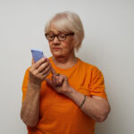 Ältere Frau in orangenem T-Shirt vor einer hellen Wand stehend, die auf ihrem Smartphone tippt