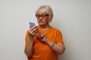 Ältere Frau in orangenem T-Shirt vor einer hellen Wand stehend, die auf ihrem Smartphone tippt