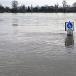 Hochwassergebiet, knapp über der Wasserfläche sieht man ein Verkehrsschild, im Hintergrund Bäume und eine Stadtsilhouette