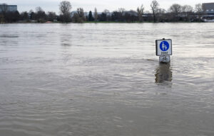 Hochwassergebiet, knapp über der Wasserfläche sieht man ein Verkehrsschild, im Hintergrund Bäume und eine Stadtsilhouette