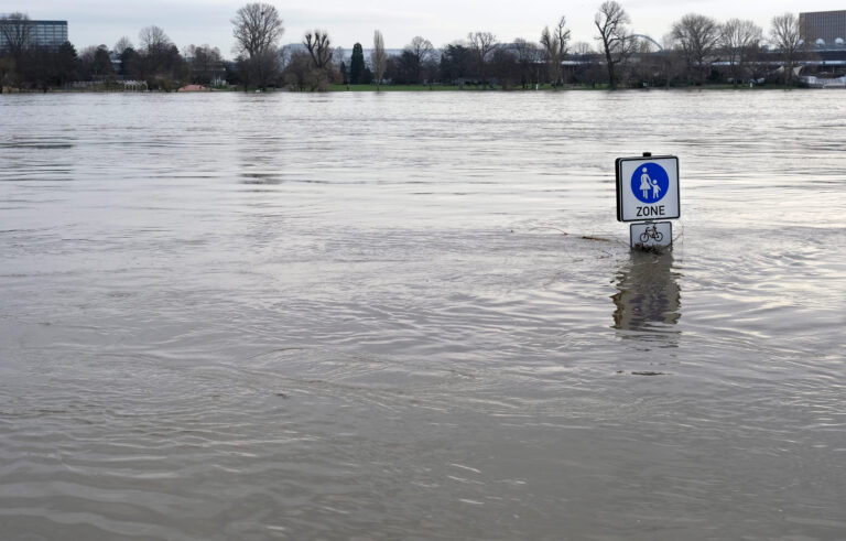 Hochwassergebiet, knapp über der Wasserfläche sieht man ein Verkehrsschild, im Hintergrund Bäume und eine Stadtsilhouette