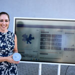 Lächelnde Frau im geblümten Sommerkleid vor einem großen Monitor.
