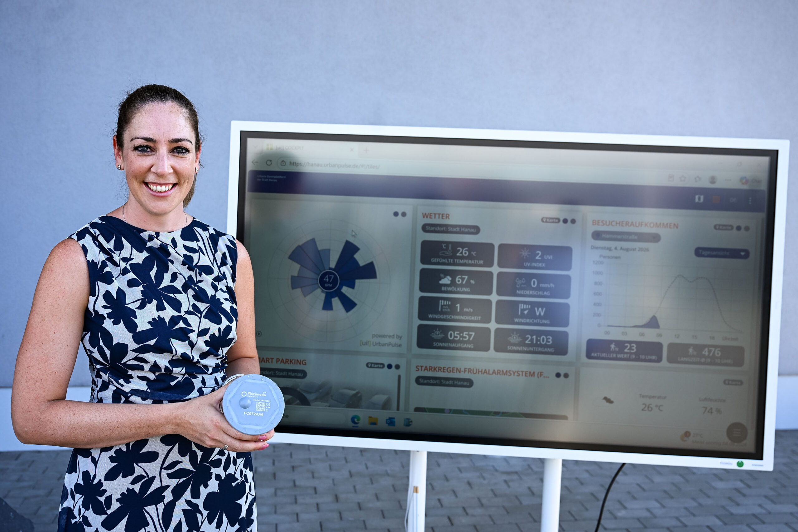 Lächelnde Frau im geblümten Sommerkleid vor einem großen Monitor.