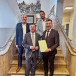 Gruppenfoto mit vier anzugtragenden Männern auf einer Treppe, einer präsentiert eine Urkunde