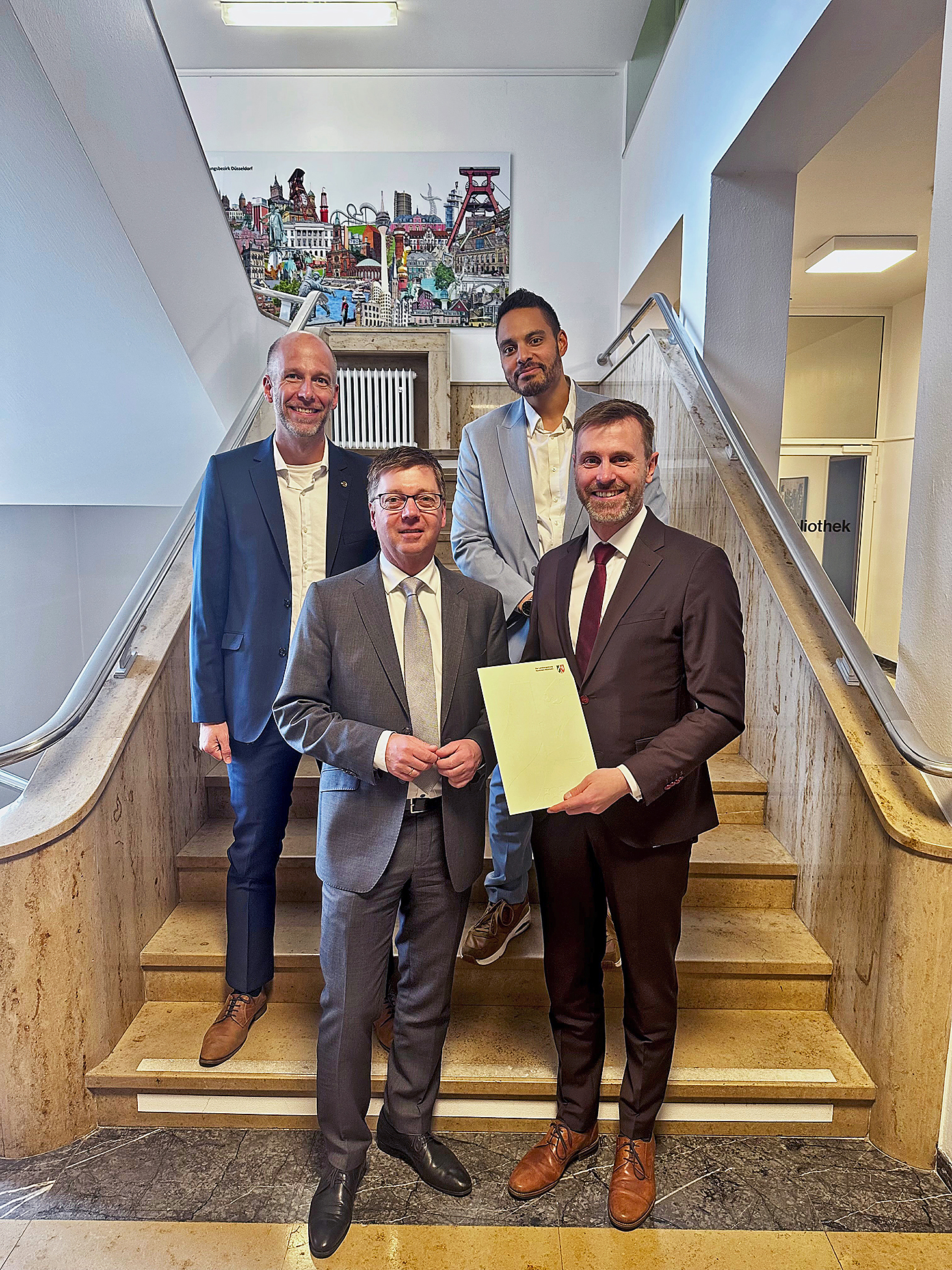 Gruppenfoto mit vier anzugtragenden Männern auf einer Treppe, einer präsentiert eine Urkunde
