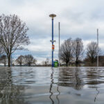 Überschwemmtes Gebiet, im Vordergrund eine Wasserfläche, hinten Bäume und Verkehrszeichen