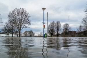 Überschwemmtes Gebiet, im Vordergrund eine Wasserfläche, hinten Bäume und Verkehrszeichen