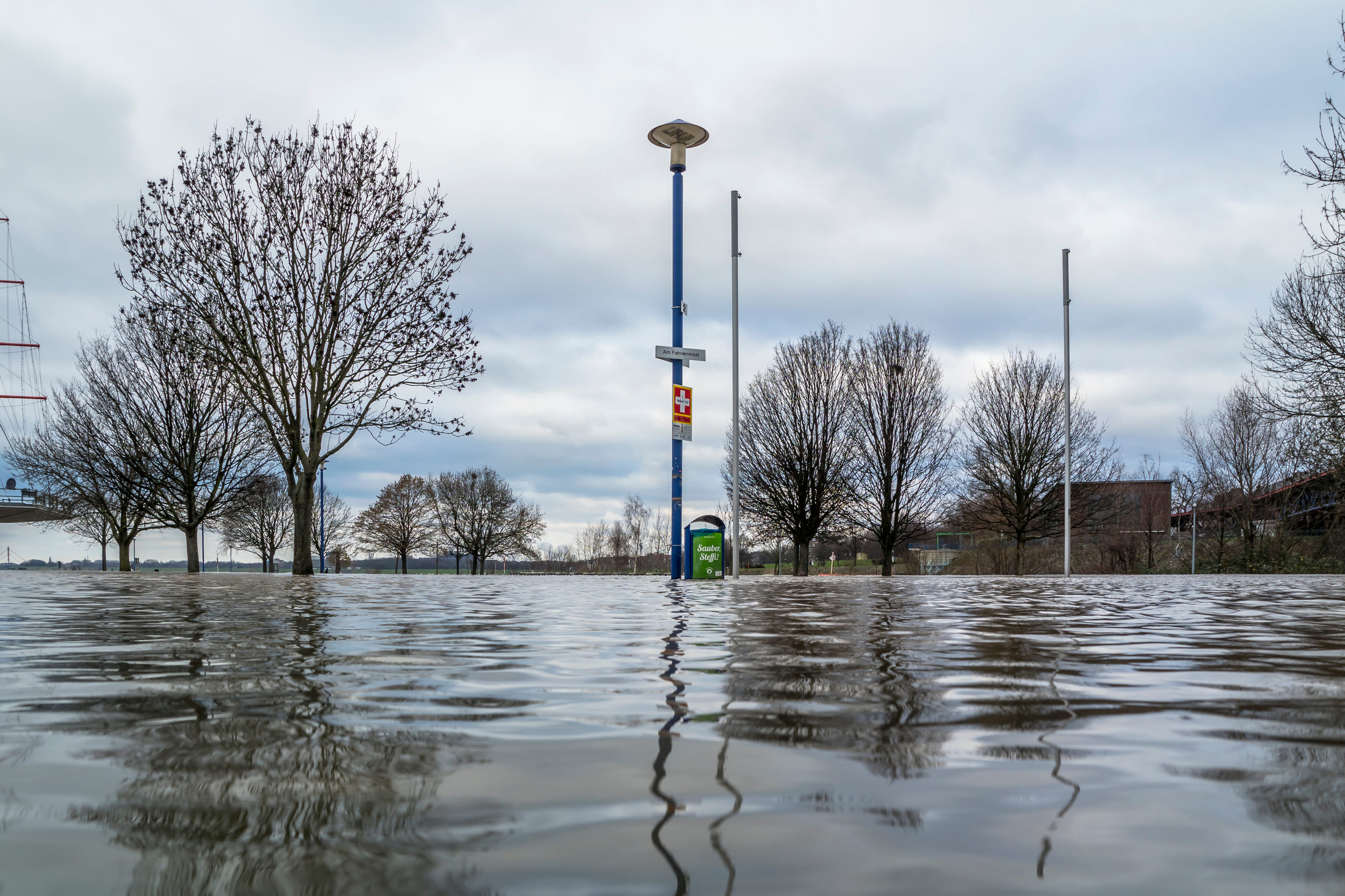 Überschwemmtes Gebiet, im Vordergrund eine Wasserfläche, hinten Bäume und Verkehrszeichen