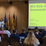 Abgedunkelter Saal mit Holzvertäfelung, Blick über Zushcauerköpfe aufs Podium, wo man eine grüße Projektion und verschiedene Flaggen sieht.