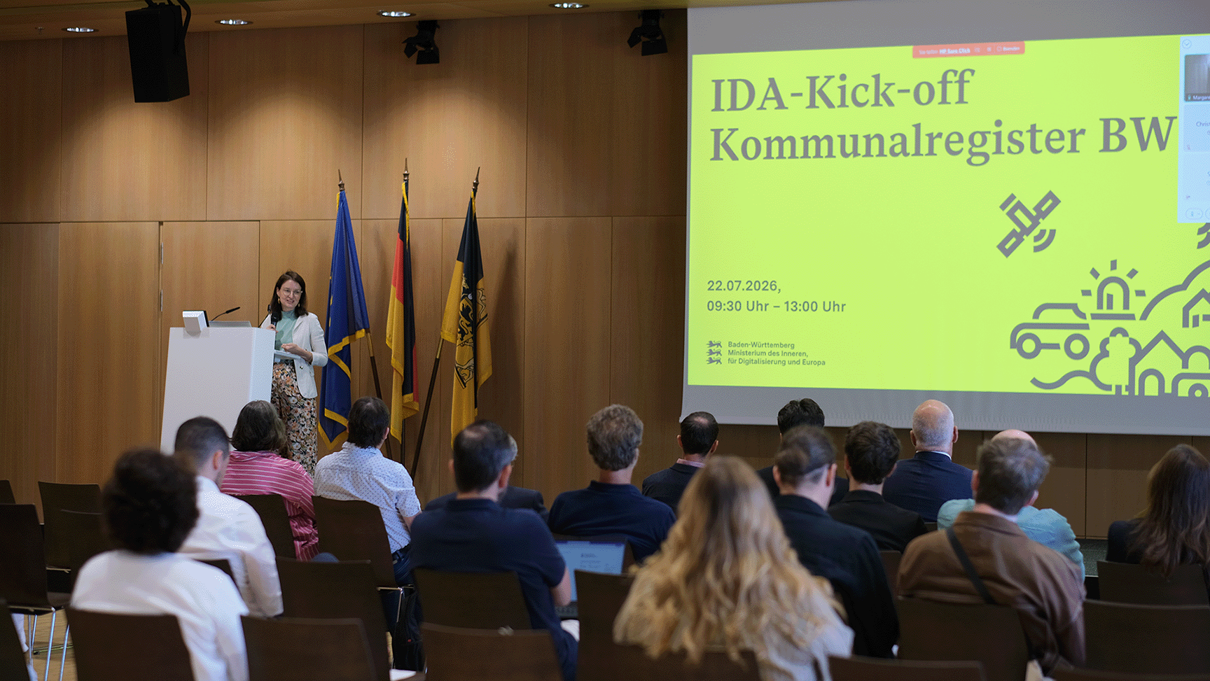 Abgedunkelter Saal mit Holzvertäfelung, Blick über Zushcauerköpfe aufs Podium, wo man eine grüße Projektion und verschiedene Flaggen sieht.
