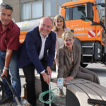 Drei Menschen mit Spaten, im Hintergrund ein orangefarbenes Baufahrzeug