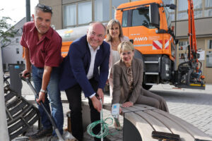 Drei Menschen mit Spaten, im Hintergrund ein orangefarbenes Baufahrzeug