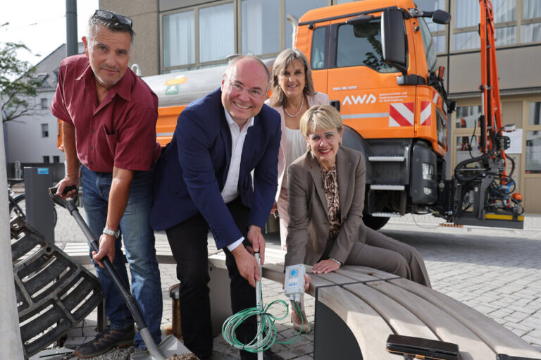 Drei Menschen mit Spaten, im Hintergrund ein orangefarbenes Baufahrzeug