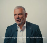 Screenshot: Mann im dunklen Anzug vor professionell ausgeleuchteter heller Wand